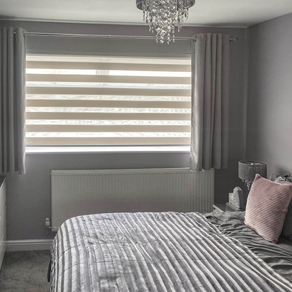 Zebra roller blind with alternating opaque and sheer horizontal bands lowered across a wide window, flanked by pleated grey curtains at the sides; filtering soft daylight into a grey-toned bedroom.