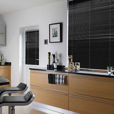 Black horizontal blinds cover a large window and smaller door window, fully lowered and closed, filtering light in a modern kitchen with light wood cabinets, black countertop and bar stools.