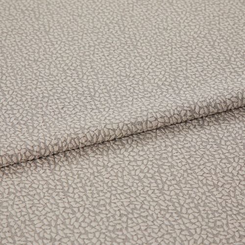 Beige jacquard-like upholstery fabric, folded over itself, displaying a dense small pebble motif; close-up studio-lit view highlighting subtle sheen, woven texture, and gentle drape.