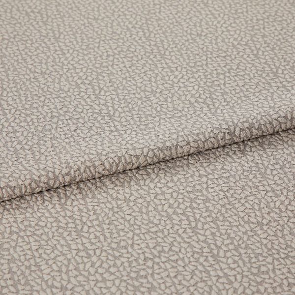 Beige jacquard-like upholstery fabric, folded over itself, displaying a dense small pebble motif; close-up studio-lit view highlighting subtle sheen, woven texture, and gentle drape.