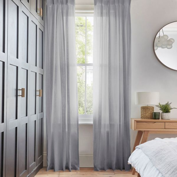 Sheer gray floor-length curtains hung either side of a sash window, filtering soft daylight and parted to show foliage outside; bedroom with dark paneled wardrobe and bedside table.