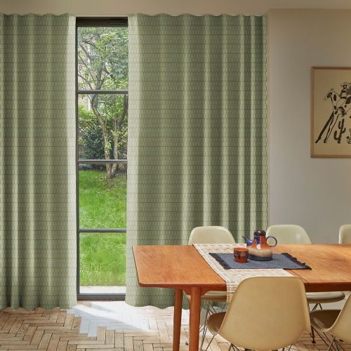 Green patterned curtains, floor-length on each side of a tall three-pane window, hanging in soft folds, partially open and filtering daylight; dining room with wooden table, chairs, garden view.