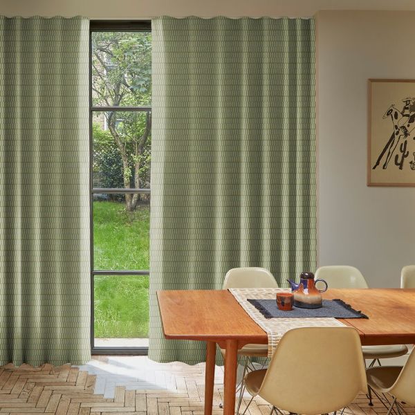 Green patterned curtains, floor-length on each side of a tall three-pane window, hanging in soft folds, partially open and filtering daylight; dining room with wooden table, chairs, garden view.