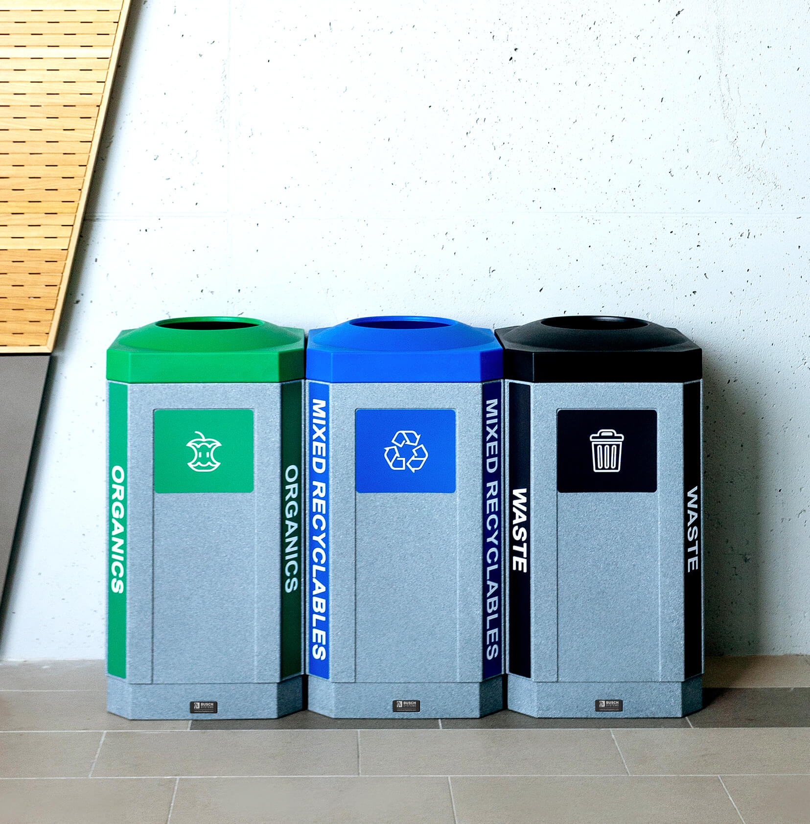 durable plastic waste recycling and organics bins with colored lids and labels sitting against a wall at a high school