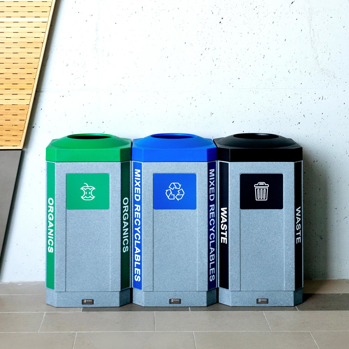 durable plastic waste recycling and organics bins with colored lids and labels sitting against a wall at a high school