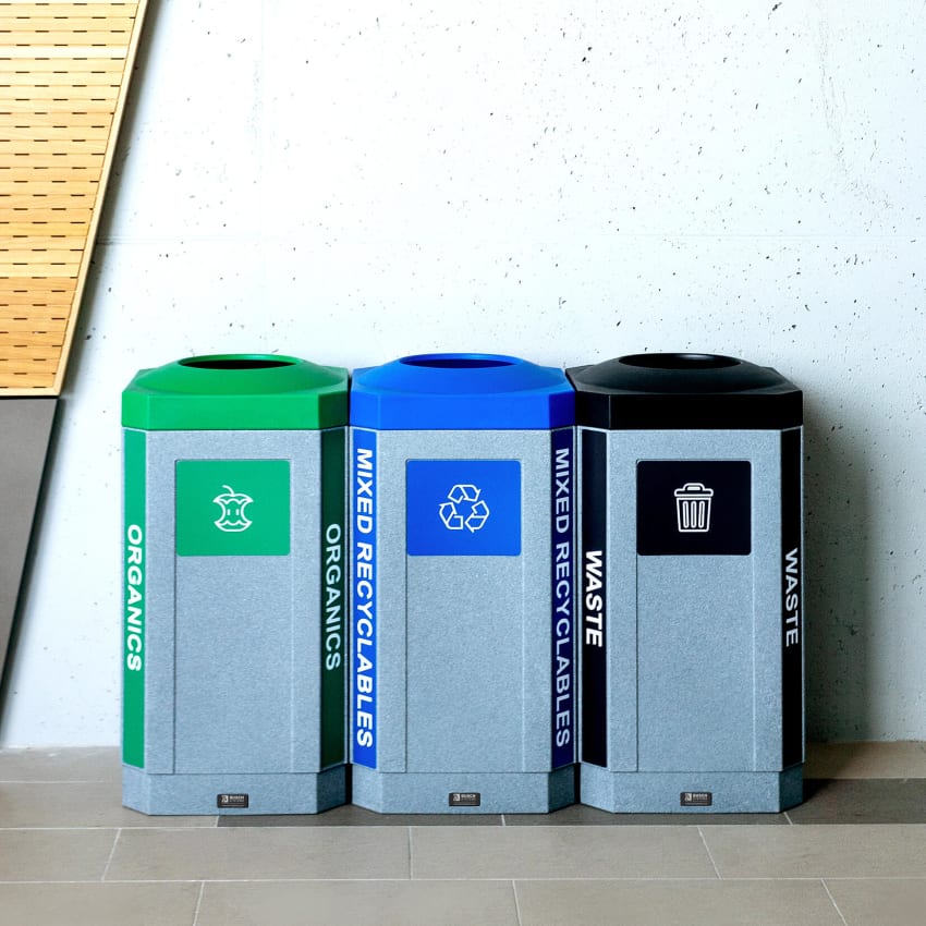 durable plastic waste recycling and organics bins with colored lids and labels sitting against a wall at a high school