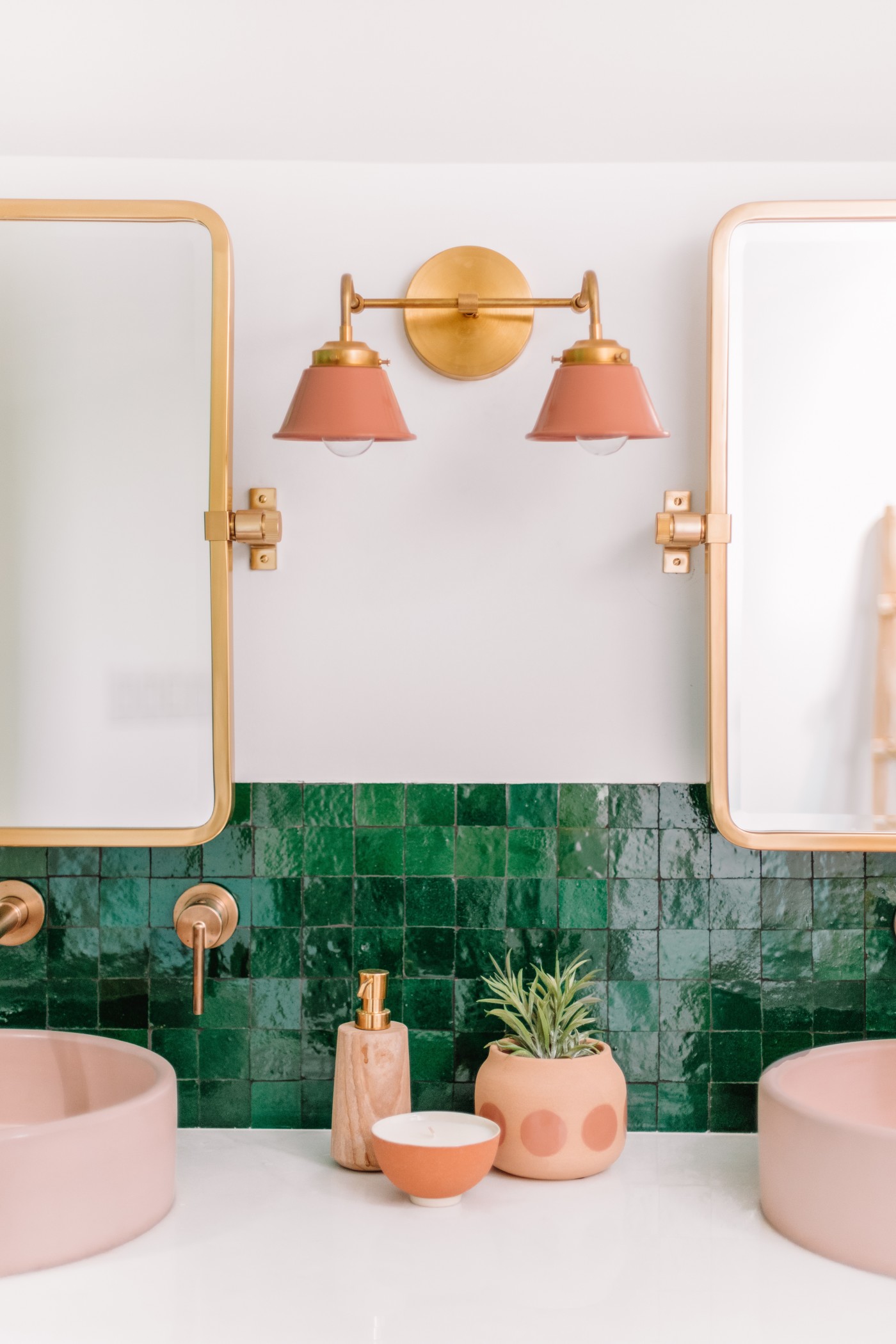 a bathroom with two pink sinks, 2 gold mirrors and a green tile backsplash.