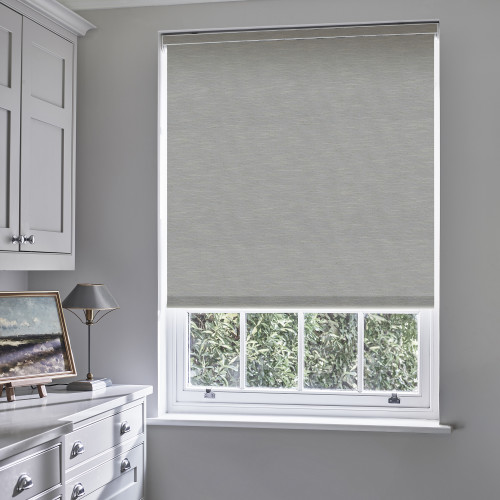 Grey roller blind, lowered over a sash window to cover the upper panes and filter daylight; pale-grey room with a dresser, lamp and framed painting, view of dense green shrubs outside.