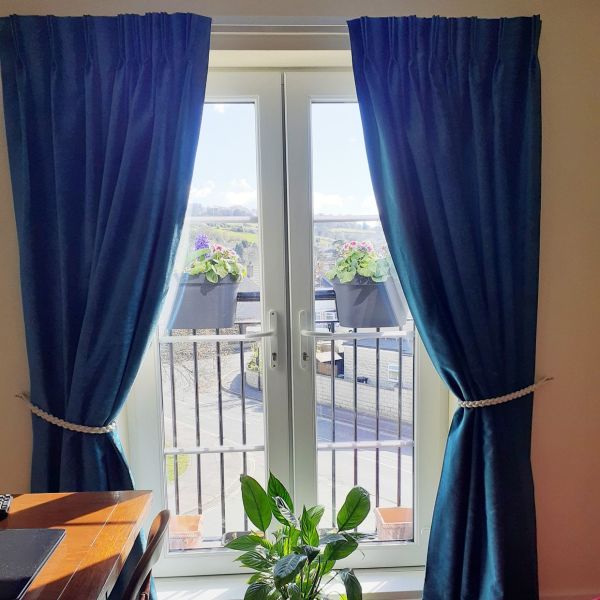 Teal pleated curtains tied back with braided ropes, parted to reveal double glass doors and a balcony with flower boxes; bright daylight fills a living room with a windowsill plant.