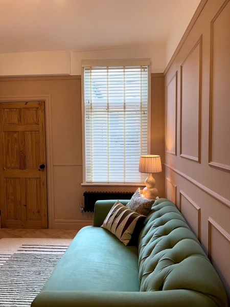 White horizontal Venetian blinds covering a tall sash window, closed and diffusing daylight; set above a black radiator in a warmly lit sitting room with green tufted sofa and lamp.