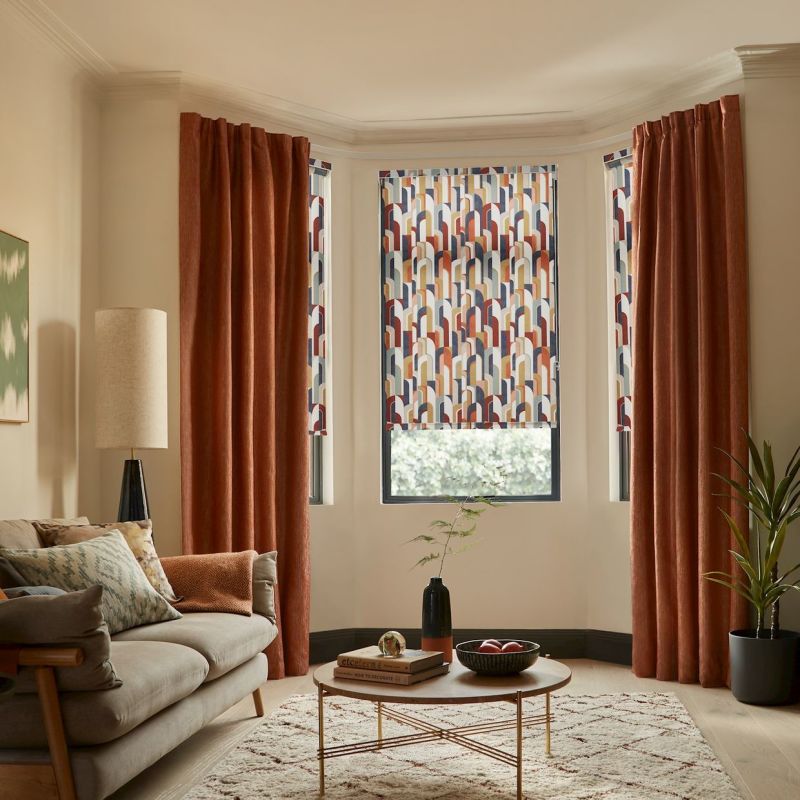 Patterned roller blind and rust velvet curtains in a bay window: blind lowered with geometric arches, curtains drawn to sides; warm living room with sofa, coffee table, rug.