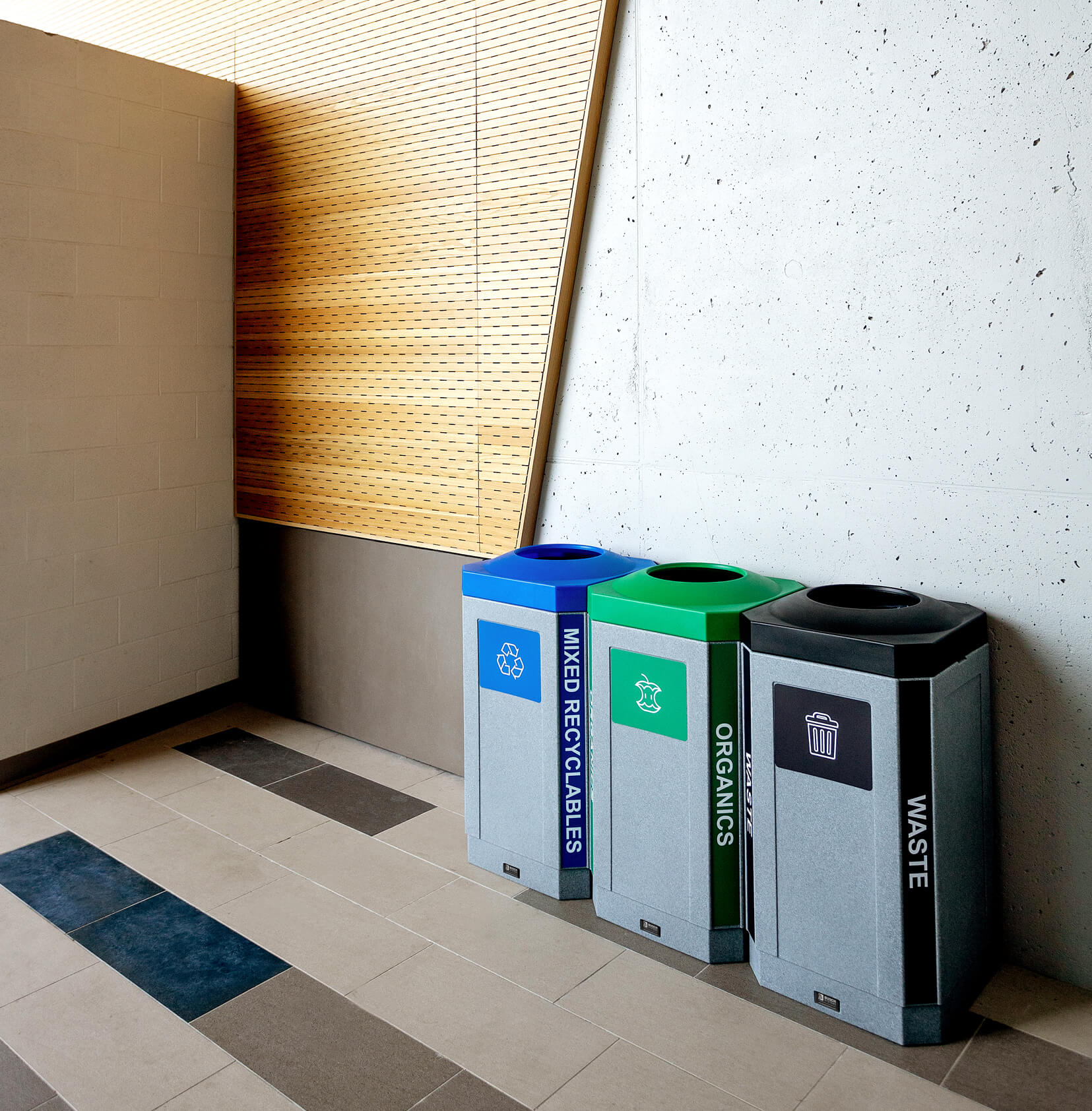 grey plastic triple waste recycling and organics containers with colored lids and labels in a modern college campus
