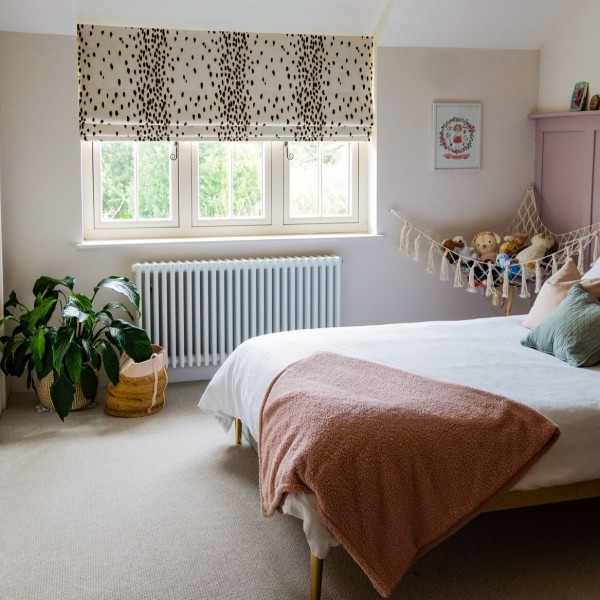 Cream Roman blind with irregular dark spots lowered across a three-pane window, partially filtering daylight in a pastel bedroom with bed, pink throw, potted plant and stuffed-animal hammock.