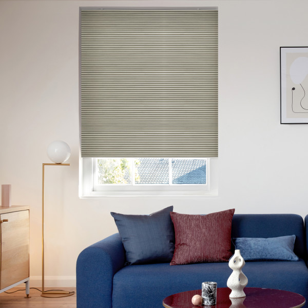 Beige narrow‑pleated blind, lowered to cover most of the window with a bottom gap, filtering daylight. Bright living room with blue sofa, cushions, floor lamp and framed art; rooftop beyond.