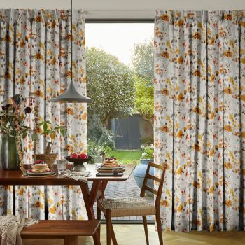 Pleated full-length floral curtains (white with yellowâ€‘orange flowers) are parted to reveal a sliding glass door, filtering soft daylight over a breakfast table and wooden chairs overlooking a sunlit garden.
