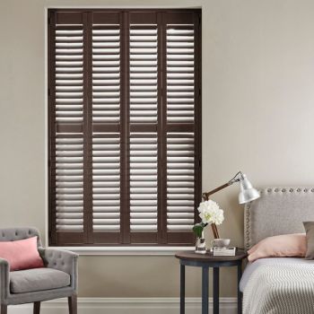 Dark wooden plantation shutters—four tall louvered panels covering the window—are closed with slats slightly tilted, filtering soft daylight into a neutral-toned bedroom with bed, armchair, and bedside table.