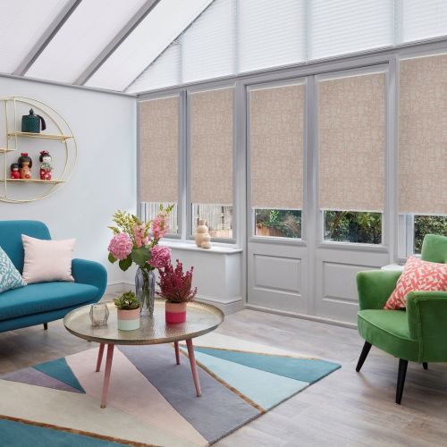Beige patterned roller blinds cover tall conservatory doors and lower windows, partially closed to filter daylight, in a bright, pastel-furnished sunroom with colorful sofa, rug, and plants.