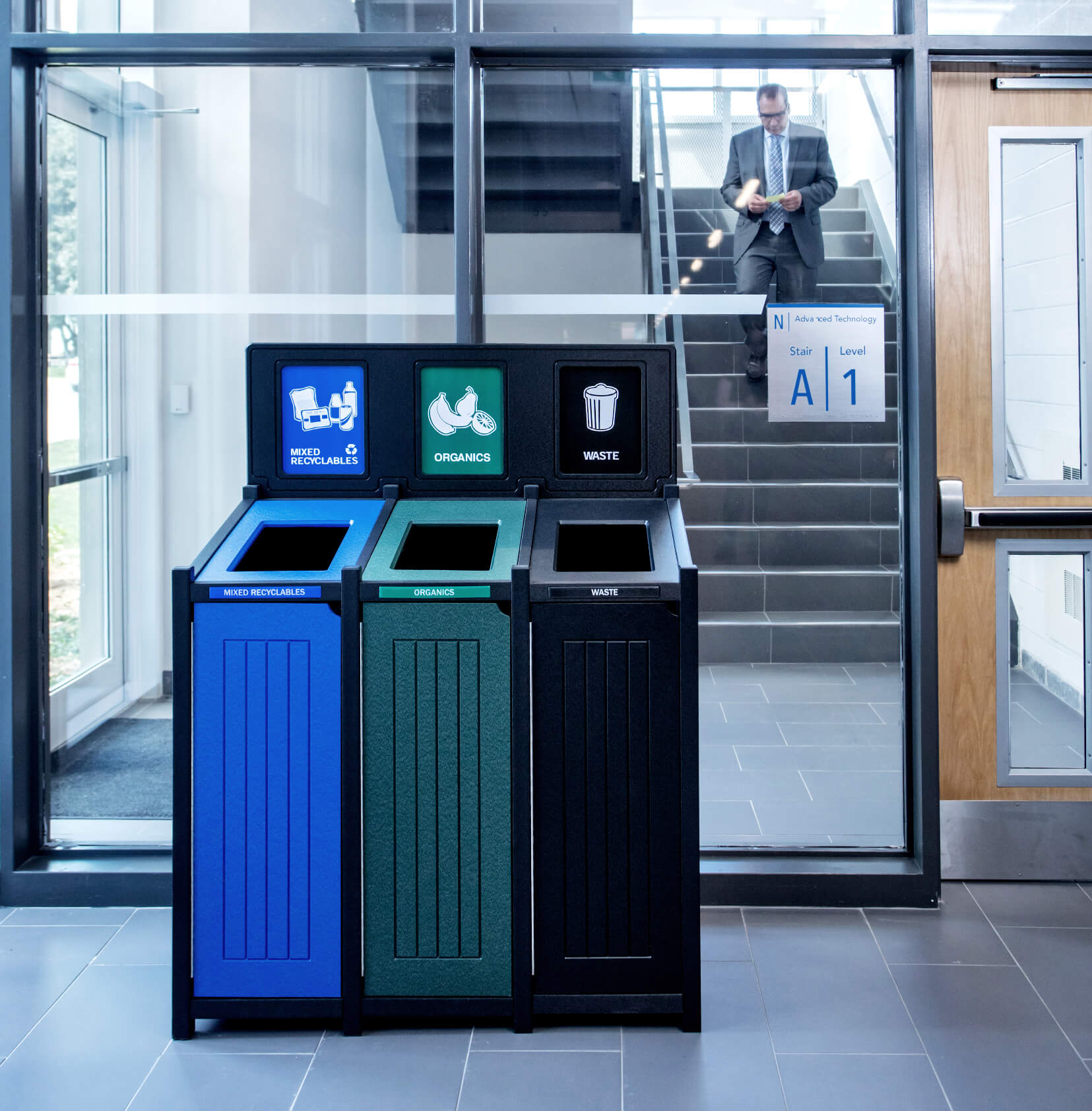 triple recycling organics and waste bin against a glass wall in a modern office with man walking down staircase