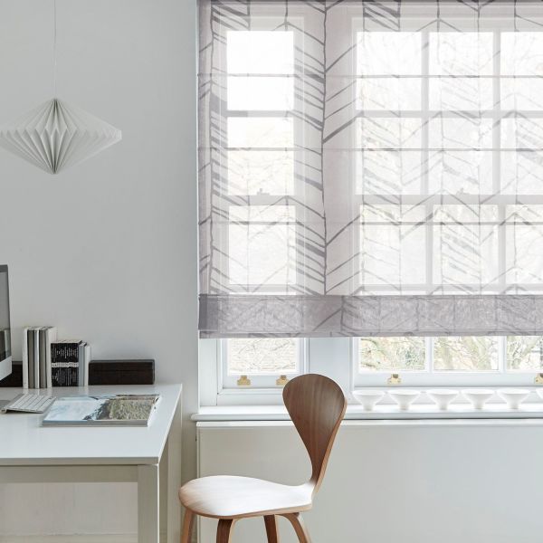 Sheer patterned Roman blind (leaf-chevron motif), lowered to mid-height and filtering soft daylight through a paneled window; minimal white home office with wooden chair, desk, pendant paper lamp, tree view.