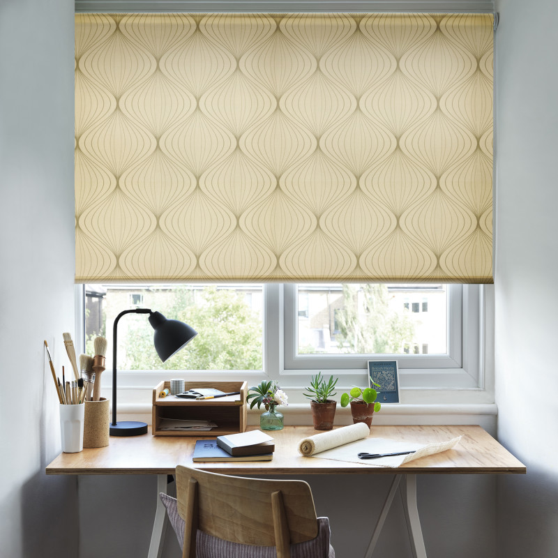 Patterned roller blind covering the upper two-thirds of a window, pale gold with curving geometric motifs — lowered to filter daylight — small wooden desk, lamp, art supplies and potted plants beneath.