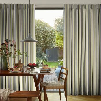 Floor-to-ceiling pleated striped curtains (gray, white, yellow) frame a central glass door, parted to reveal sunlight and a leafy backyard; foreground dining table set with pancakes, pendant lamp, wooden chairs.