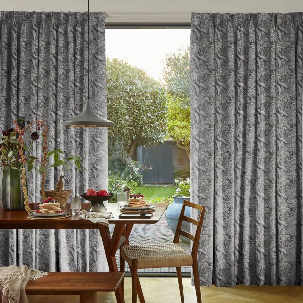 Floor-length pleated grey floral-patterned curtains, drawn apart at center, framing a sunlit garden; they filter soft light onto a wooden dining table set for breakfast with pendant lamp and chairs.