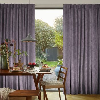 Pleated mauve floor-length curtains, drawn aside to reveal a central opening; framing and filtering soft daylight in a dining area with a wooden table and leafy garden view.