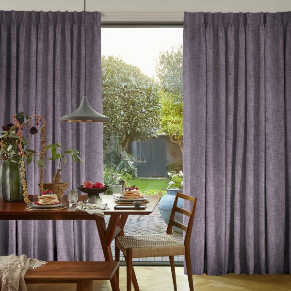 Pleated mauve floor-length curtains, drawn aside to reveal a central opening; framing and filtering soft daylight in a dining area with a wooden table and leafy garden view.