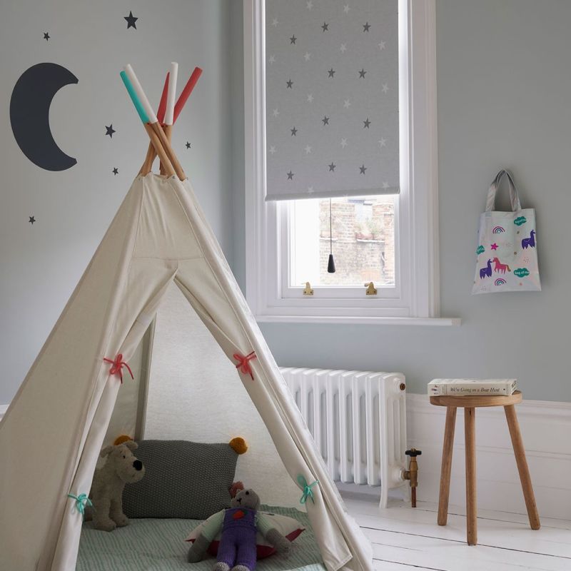 Grey roller blind with small star pattern lowered over a sash window, partially filtering daylight; canvas teepee with toys occupies foreground in a pale-grey children's bedroom, with radiator and stool.
We're Going on a Bear Hunt