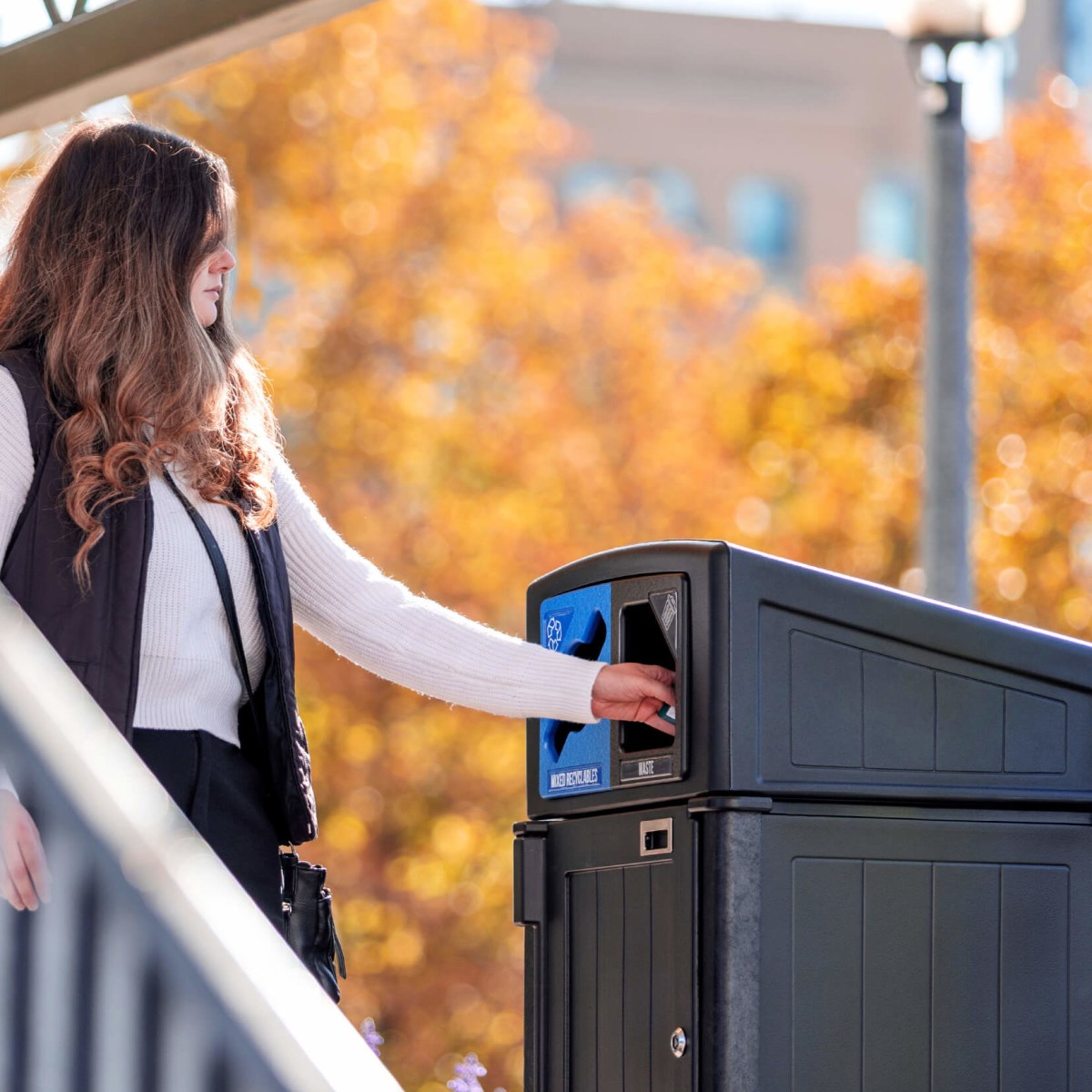 woman putting trash into black recycled plastic dual stream waste and recycling container with fall trees in the background