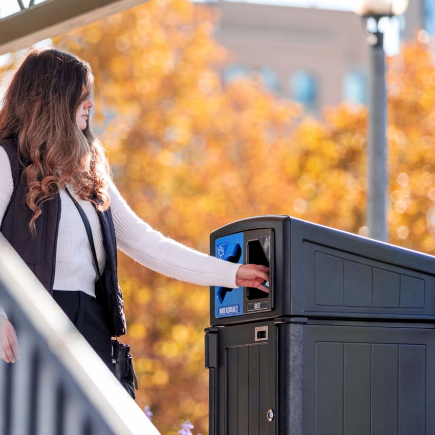 woman putting trash into black recycled plastic dual stream waste and recycling container with fall trees in the background