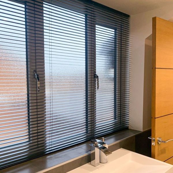 Dark horizontal Venetian blinds cover a three-panel frosted window to sill; slats slightly tilted, filtering daylight and obscuring view. Bathroom sink with chrome faucet in foreground beside wooden door.