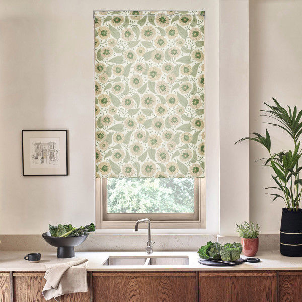 Floral-patterned roller blind covers a kitchen window, filtering light above a sink; countertop holds bowls of greens, a towel, framed sketch on wall and potted plants.