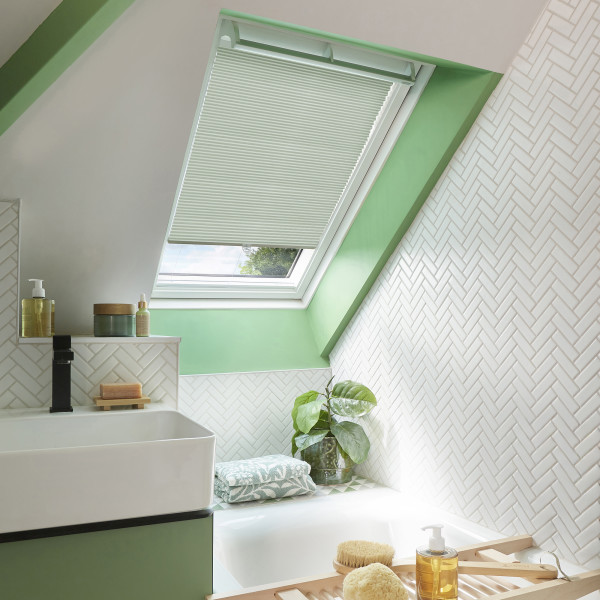 Pleated white cellular blind on an angled skylight, partially lowered and filtering daylight; sits above a green-framed attic bathroom with herringbone white tiles, bathtub, towel, potted plant, and toiletries.