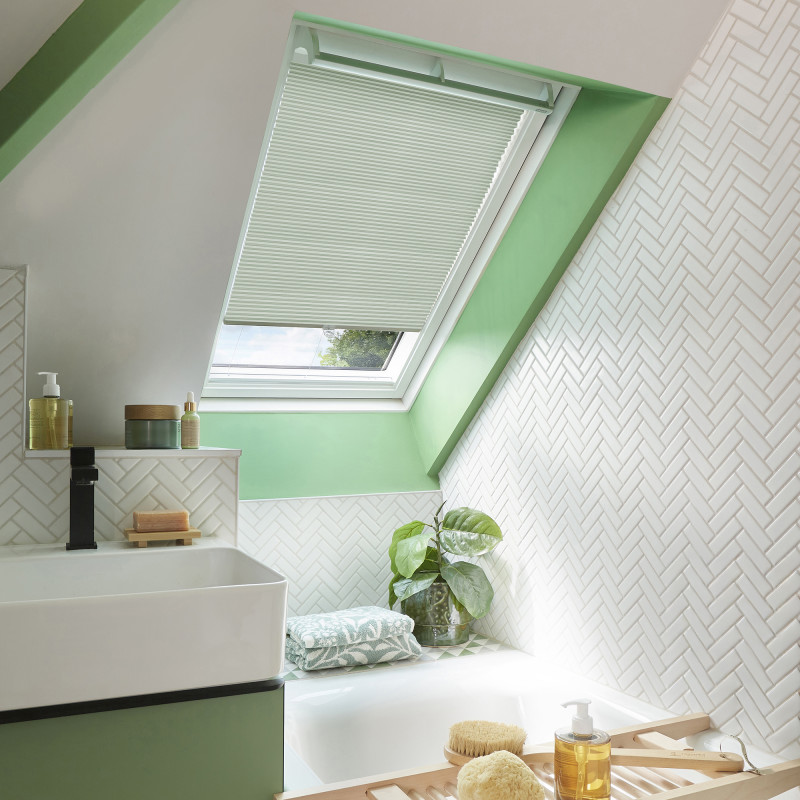 Pleated white cellular blind on an angled skylight, partially lowered and filtering daylight; sits above a green-framed attic bathroom with herringbone white tiles, bathtub, towel, potted plant, and toiletries.