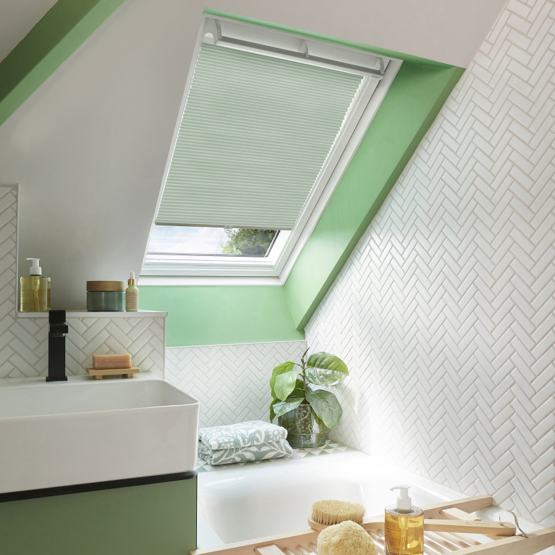 Mint-green pleated skylight blind, lowered and filtering daylight with a narrow open gap, in an angled bathroom window; white herringbone tiles, green trim, sink, tub, and potted plant nearby.