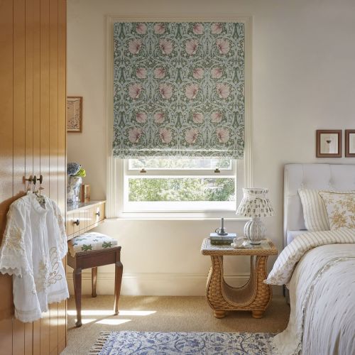 Floral-patterned Roman blind, lowered halfway, featuring green background and pink blossoms, filters soft daylight through a sash window into a cozy, softly lit bedroom with wicker bedside table.