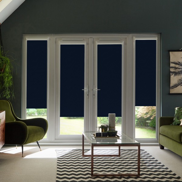 French patio doors covered by navy roller blinds partially lowered, revealing narrow strips of daylight; modern living room with green armchairs, glass coffee table on chevron rug and potted plants.