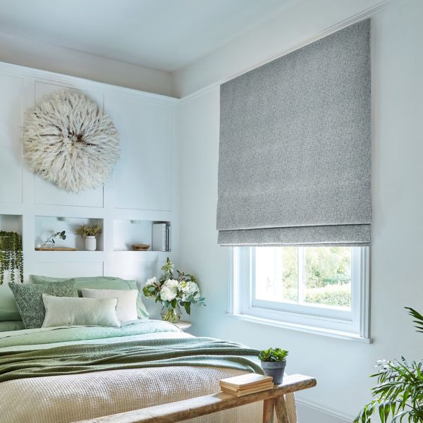 Grey textured Roman blind covering a window, lowered to block and filter light; in a brightly lit bedroom with green-accent bedding, built-in headboard niches, plants, and a feather wall hanging.