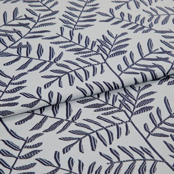 Light gray woven fabric with navy embroidered fern-like leaf pattern, folded in the center creating overlapping layers; close-up, soft diffuse lighting reveals textile texture and stitch detail.