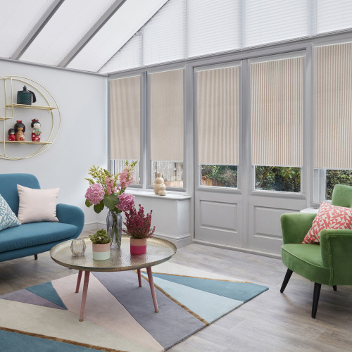 Beige pleated blinds with vertical ribbing, lowered over tall glass doors and windows to just above sills, filtering daylight; bright conservatory room with teal sofa, green armchair, pastel decor.