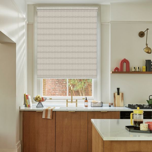 Beige textured roller blind, lowered across the window with horizontal weave; filtering soft daylight. Kitchen sink with brass faucet below, marble countertop, wooden cabinets, shelf with objects, brick courtyard outside.