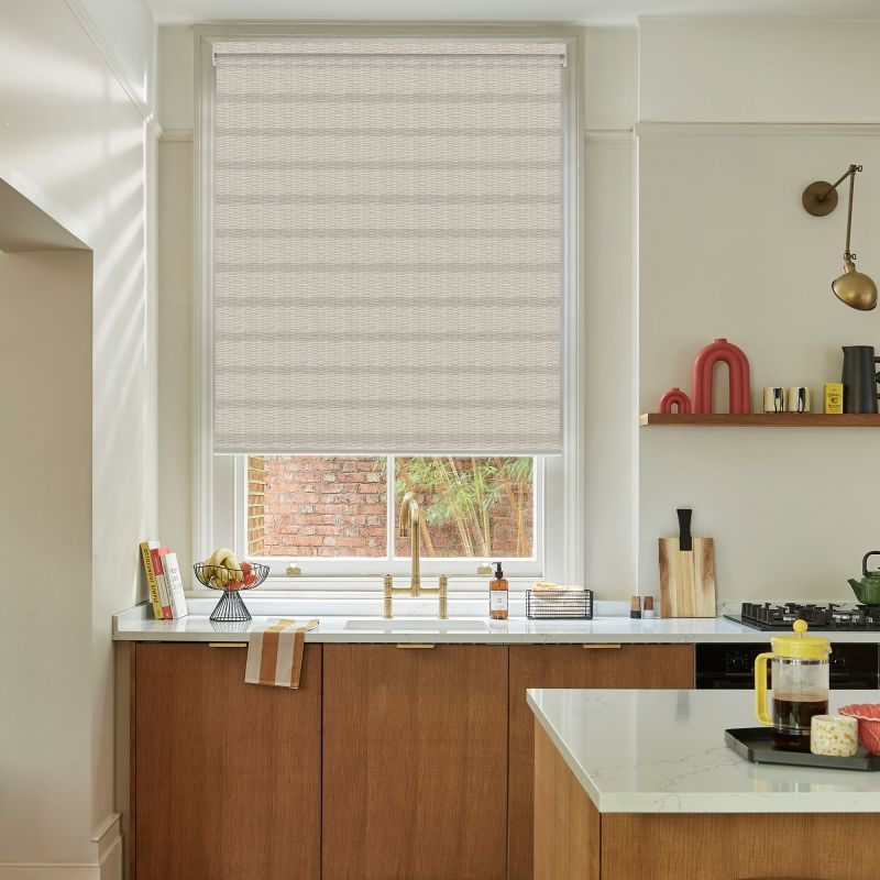 Beige textured roller blind, lowered across the window with horizontal weave; filtering soft daylight. Kitchen sink with brass faucet below, marble countertop, wooden cabinets, shelf with objects, brick courtyard outside.
