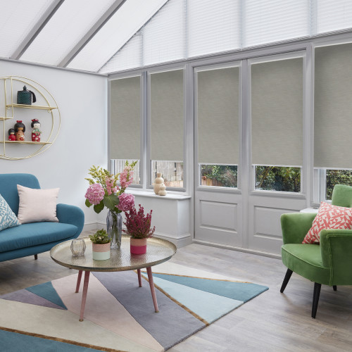 Light-gray roller blinds on tall windows and French doors, lowered to reveal narrow bottom panes and diffuse daylight; conservatory living room with teal sofa, green armchair, coffee table, flowers.