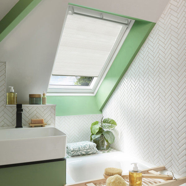 White pleated skylight blind, partially lowered over a roof window, filtering soft daylight into a bright attic bathroom framed in mint-green trim with white herringbone tiles, sink, tub, plant.