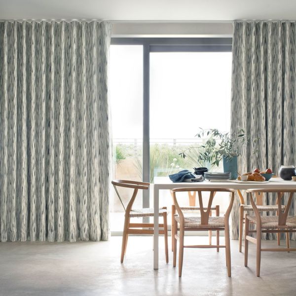 Full-length, pleated patterned curtains (greyâ€‘white vertical brushstrokes) partially drawn, framing a sliding glass door and filtering bright daylight; dining table with wooden chairs, tableware, plant, balcony view.