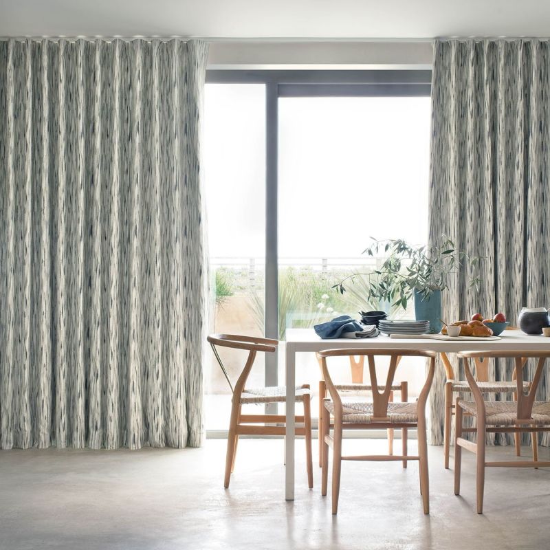 Full-length, pleated patterned curtains (greyâ€‘white vertical brushstrokes) partially drawn, framing a sliding glass door and filtering bright daylight; dining table with wooden chairs, tableware, plant, balcony view.