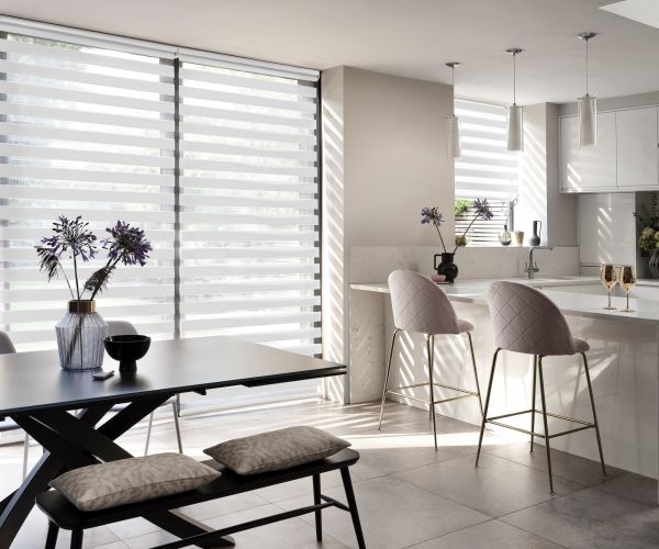 Striped dualâ€‘layer roller blinds across full-height windows, alternating sheer and opaque bands, filtering sunlight and casting horizontal shadows across a modern open-plan kitchen-dining area with marble island and bar stools.