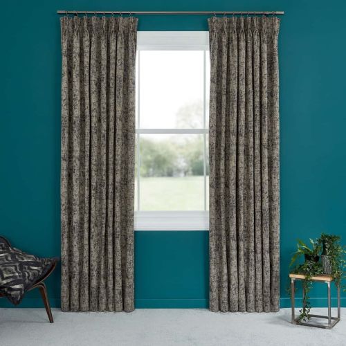 Full-length pleated gray textured curtains, on a metal rod and pulled aside, framing a white window; teal-painted wall, carpeted floor, chair left and potted plant on table right, daylight outside.