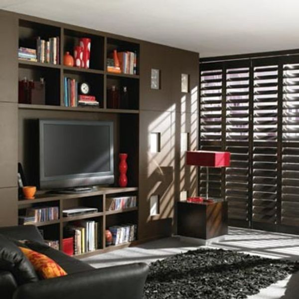 Dark wooden plantation shutters covering a floor-to-ceiling window, louvers angled to filter sunlight into striped shadows across a contemporary living room with built-in media shelving, TV, black sofa, red accents.